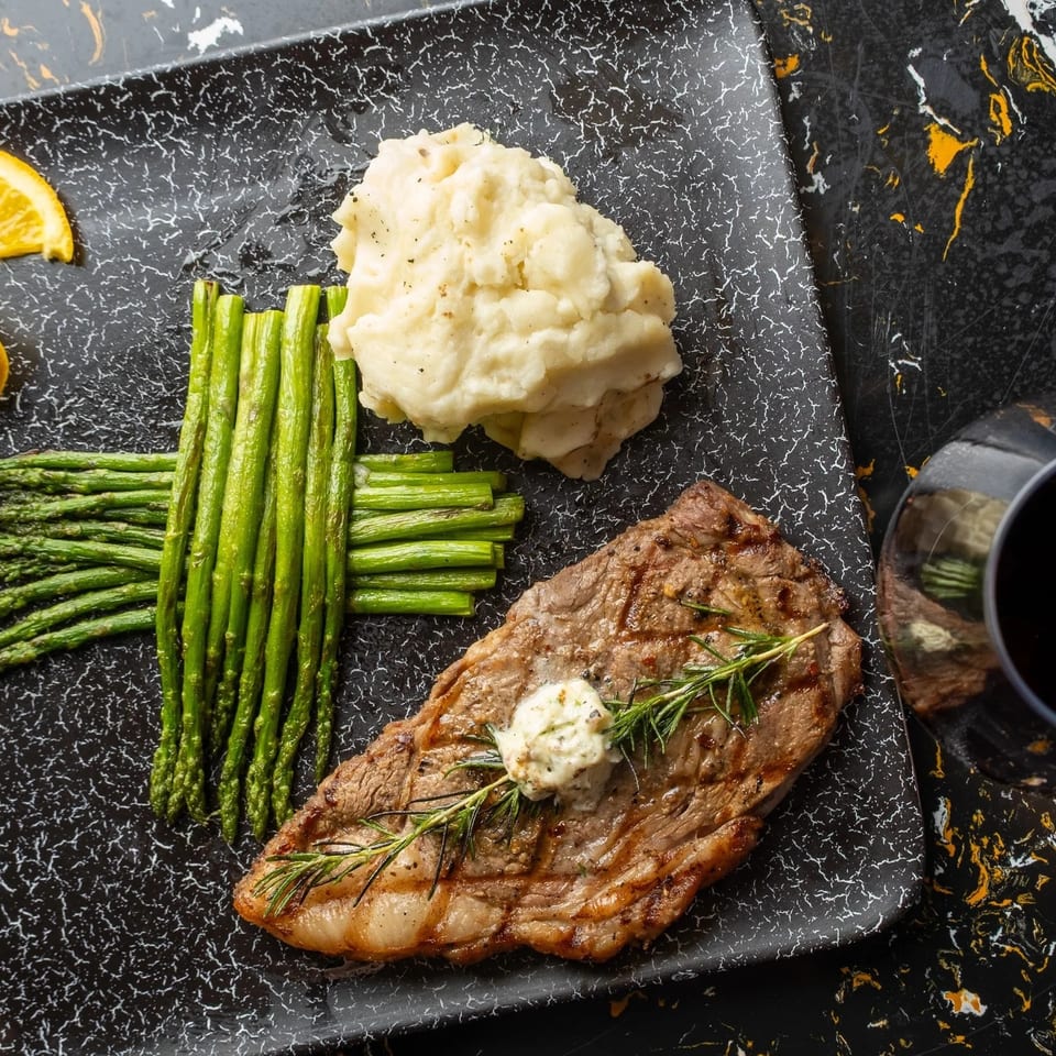 Grilled steak with herb butter, mashed potatoes, asparagus, and an orange slice on a black plate.